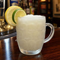 A frosted clear glass mug filled with a light yellow cocktail, garnished with two lemon slices on a dark wooden bar counter with blurred bottles in the background.