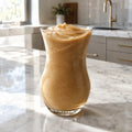 A tall, flared clear glass filled with a thick, creamy tan-brown smoothie or frozen dessert, set on a white marble kitchen countertop with a blurred kitchen background.