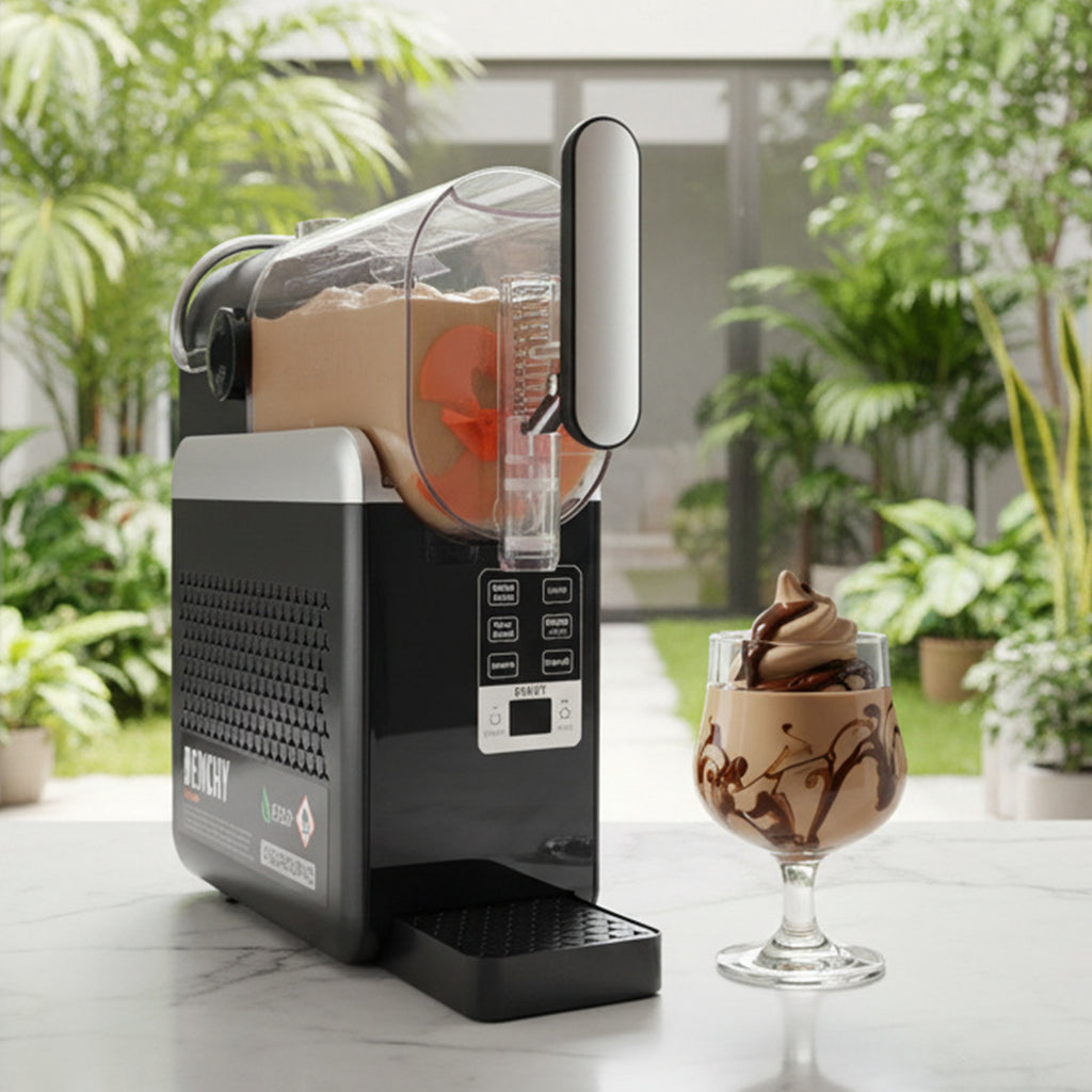 A black and silver countertop slushie maker with a clear container filled with a light brown drink, shown on a white marble surface with a glass of matching drink and a blurred indoor garden background.