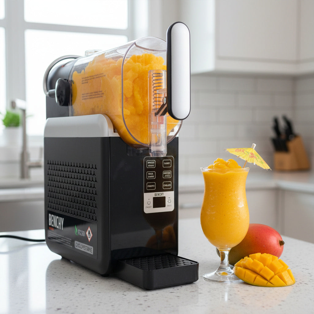 A Benchy Slushy Machine on a kitchen countertop, shown with a glass of Frozen Skinny Mango Daiquiri VINKA Slushy Cocktail Kit (2L), mango fruit, and a small umbrella in a bright kitchen setting.