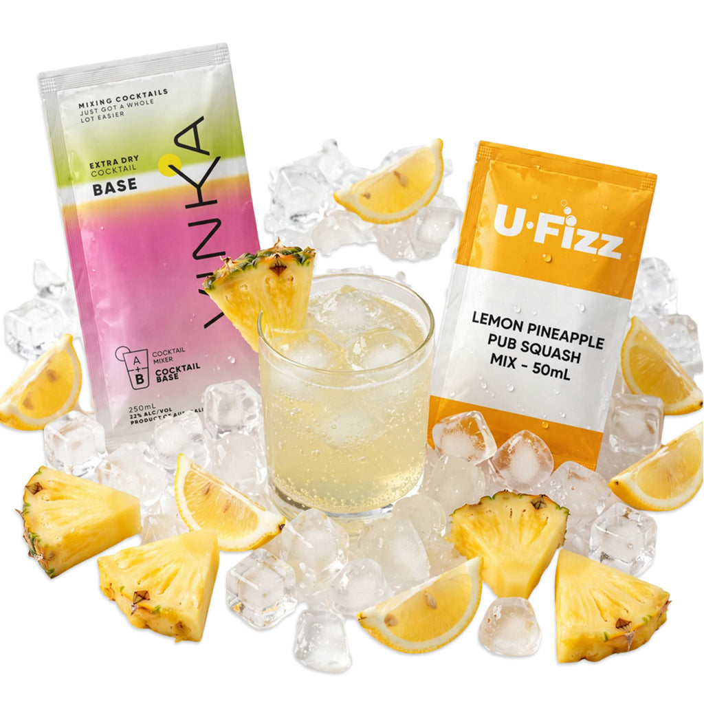 Two cocktail mix packets, one pink-to-yellow gradient labeled 'VINKA' and one orange-and-white labeled 'U-Fizz Lemon Pineapple Pub Squash Mix - 50ml', arranged around a glass of pale yellow iced drink with pineapple slices and ice cubes on a white background