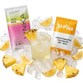 Two cocktail mix packets, one pink-to-yellow gradient labeled 'VINKA' and one orange-and-white labeled 'U-Fizz Lemon Pineapple Pub Squash Mix - 50ml', arranged around a glass of pale yellow iced drink with pineapple slices and ice cubes on a white background