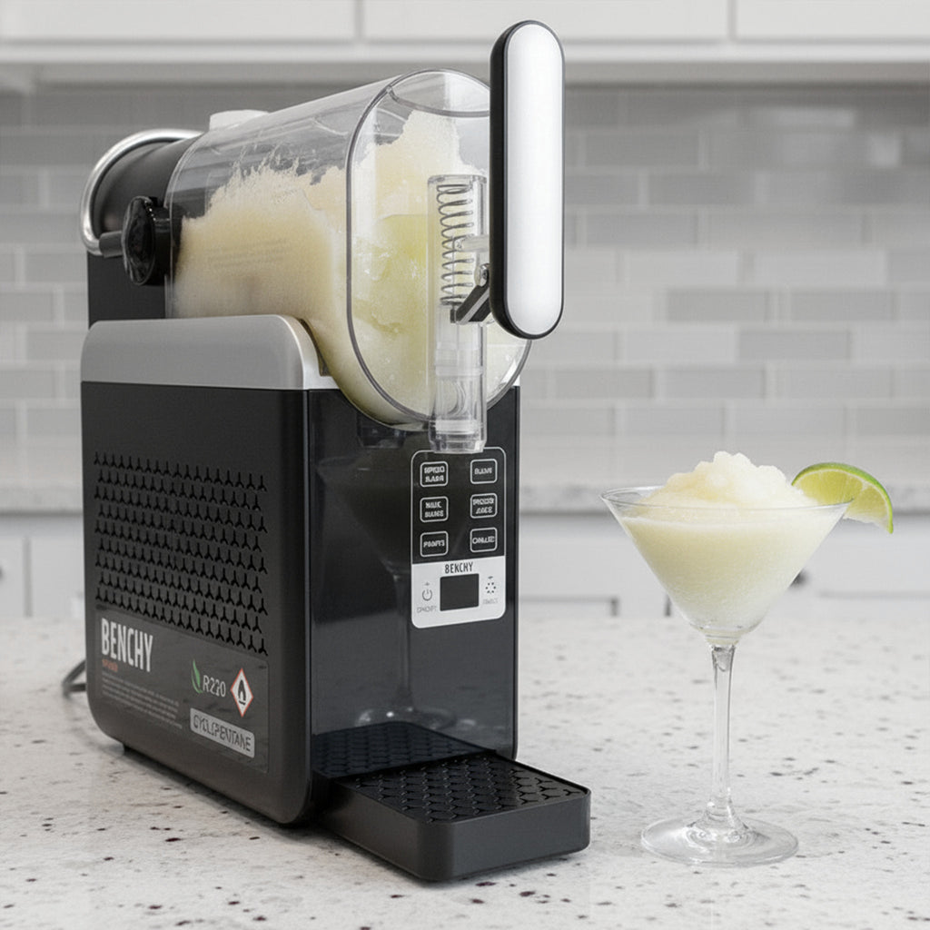 A BENCHY ice machine in black and gray sits on a kitchen counter with a glass of pale yellow slushie and a lime wedge beside it. The background shows a light gray subway tile wall.