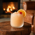 Frozen Amaretto Sour Slushy beside a fireplace. Garnished with a slice of orange and a maraschino cherry.