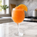 An orange frozen drink in a stemmed glass on a marble countertop, garnished with an orange slice, with a blurred kitchen background.
