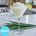 A margarita-style drink filled with Frozen Skinny Margarita VINKA Slushy Cocktail Kit (2L), garnished with a lime wedge, set on a marble countertop with aloe leaves in the foreground. A blue circle in the lower left reads “SKINNY.