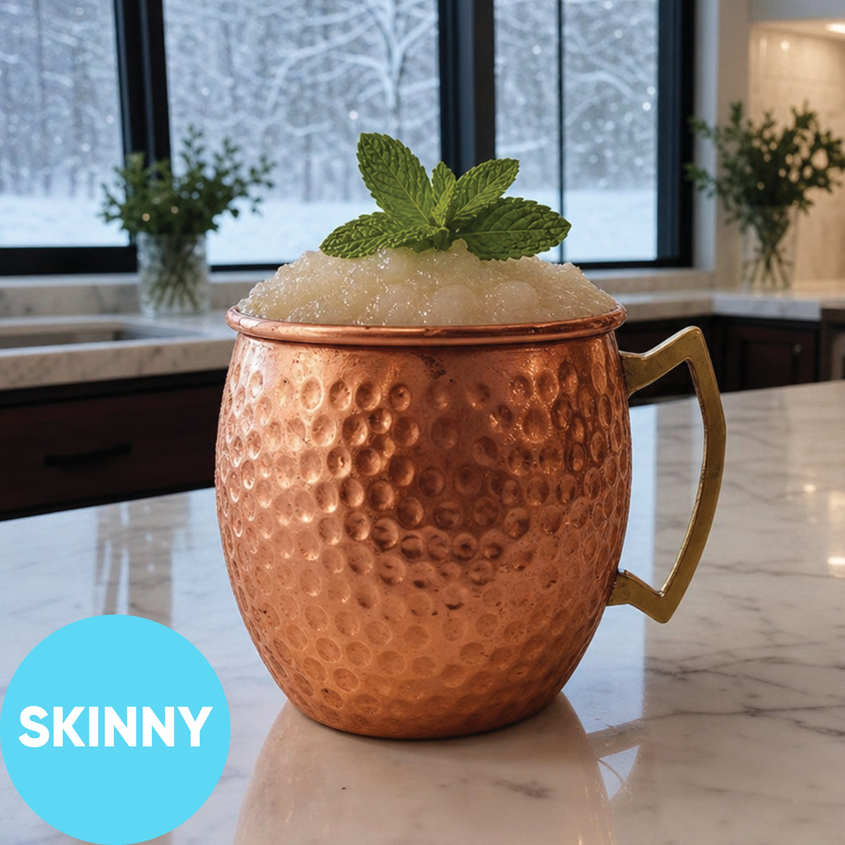 A hammered copper Moscow mule mug with a gold-toned handle, filled with Frozen Skinny Moscow Mule VINKA Slushy Cocktail Kit (2L) and mint leaves, sitting on a marble countertop in a kitchen setting. A blue circular badge in the lower left corner reads 'SKINNY'.