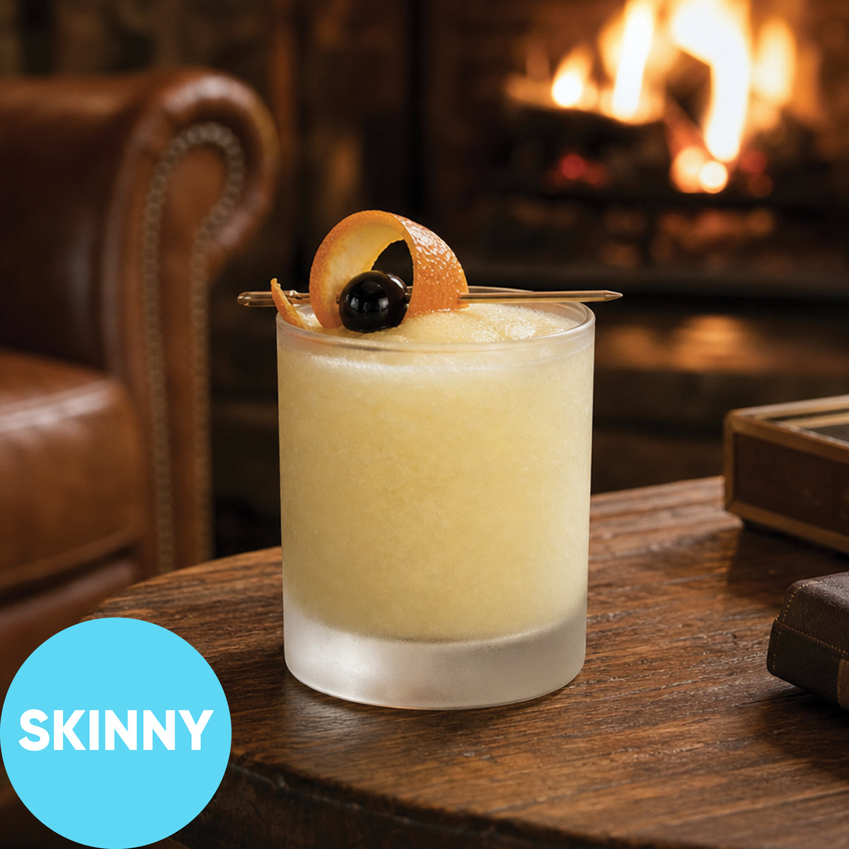 A frosted tumbler filled with Frozen Skinny Whiskey Sour VINKA Slushy Cocktail Kit (2L) sits on a wooden table in a cozy indoor setting with a fireplace and leather chair in the background. A blue circular badge in the lower left reads 'SKINNY'.