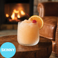 A frosted clear cocktail glass filled with Frozen Skinny Amaretto Sour VINKA Slushy Cocktail Kit (2L), garnished with an orange slice and a red cherry, set on a wooden table in front of a cozy fireplace. A blue circle in the lower left reads 'SKINNY'.