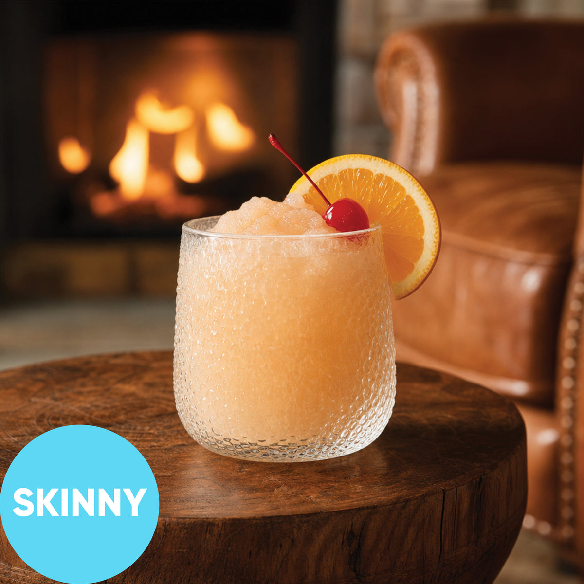 A frosted clear cocktail glass filled with Frozen Skinny Amaretto Sour VINKA Slushy Cocktail Kit (2L), garnished with an orange slice and a red cherry, set on a wooden table in front of a cozy fireplace. A blue circle in the lower left reads 'SKINNY'.