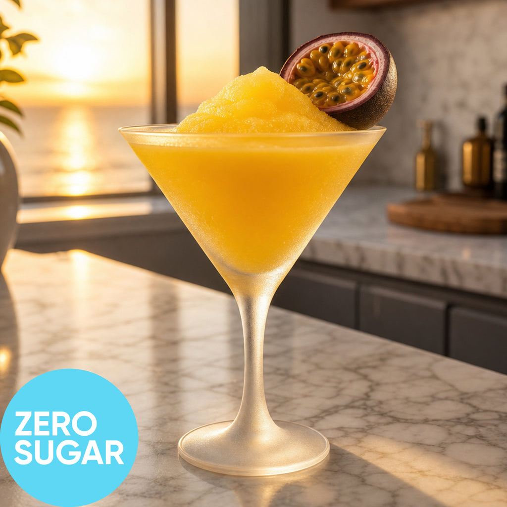 A Frozen Zero Sugar Pornstar Martini VINKA Slushy Cocktail Kit (2L) in a clear martini glass, garnished with a halved passion fruit, on a marble countertop with a sunset ocean view in the background. A blue circle in the lower left reads 'ZERO SUGAR'.