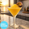 A Frozen Zero Sugar Pornstar Martini VINKA Slushy Cocktail Kit (2L) in a clear martini glass, garnished with a halved passion fruit, on a marble countertop with a sunset ocean view in the background. A blue circle in the lower left reads 'ZERO SUGAR'.