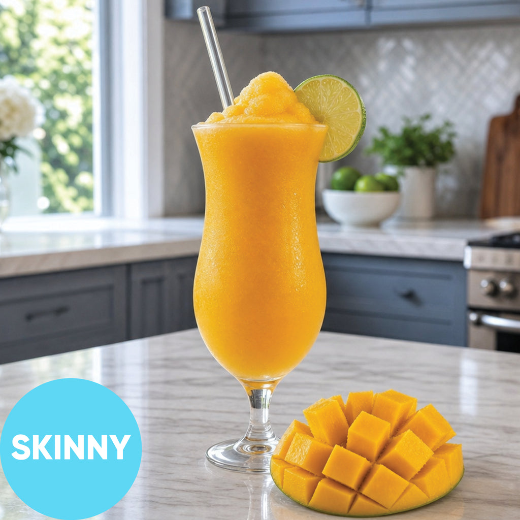 A tall glass of Frozen Skinny Mango Daiquiri VINKA Slushy Cocktail Kit (2L) with a lime wedge garnish and a straw, set on a kitchen counter next to a sliced mango. A blue circle in the lower left says 'SKINNY'.