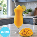 A tall glass of Frozen Skinny Mango Daiquiri VINKA Slushy Cocktail Kit (2L) with a lime wedge garnish and a straw, set on a kitchen counter next to a sliced mango. A blue circle in the lower left says 'SKINNY'.