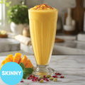 A tall clear glass filled with Frozen Skinny Hard Mango Lassi VINKA Slushy Cocktail Kit (2L), topped with saffron threads, set on a marble surface with mango slices, mint, and rose petals. A blue circle in the lower left says SKINNY.