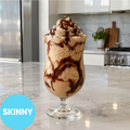 A tall glass filled with a light tan creamy dessert, topped with whipped cream and chocolate syrup, sitting on a marble kitchen counter. A blue circle in the lower left says 'SKINNY'.