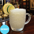 A frosted clear glass mug filled with Frozen Skinny Hard Lemon-Lime Pub Squash VINKA Slushy Kit (2L), garnished with two lime slices on the rim, set on a wooden bar counter with a blurred bar background. A blue circular badge in the lower left reads 'SKINNY'.