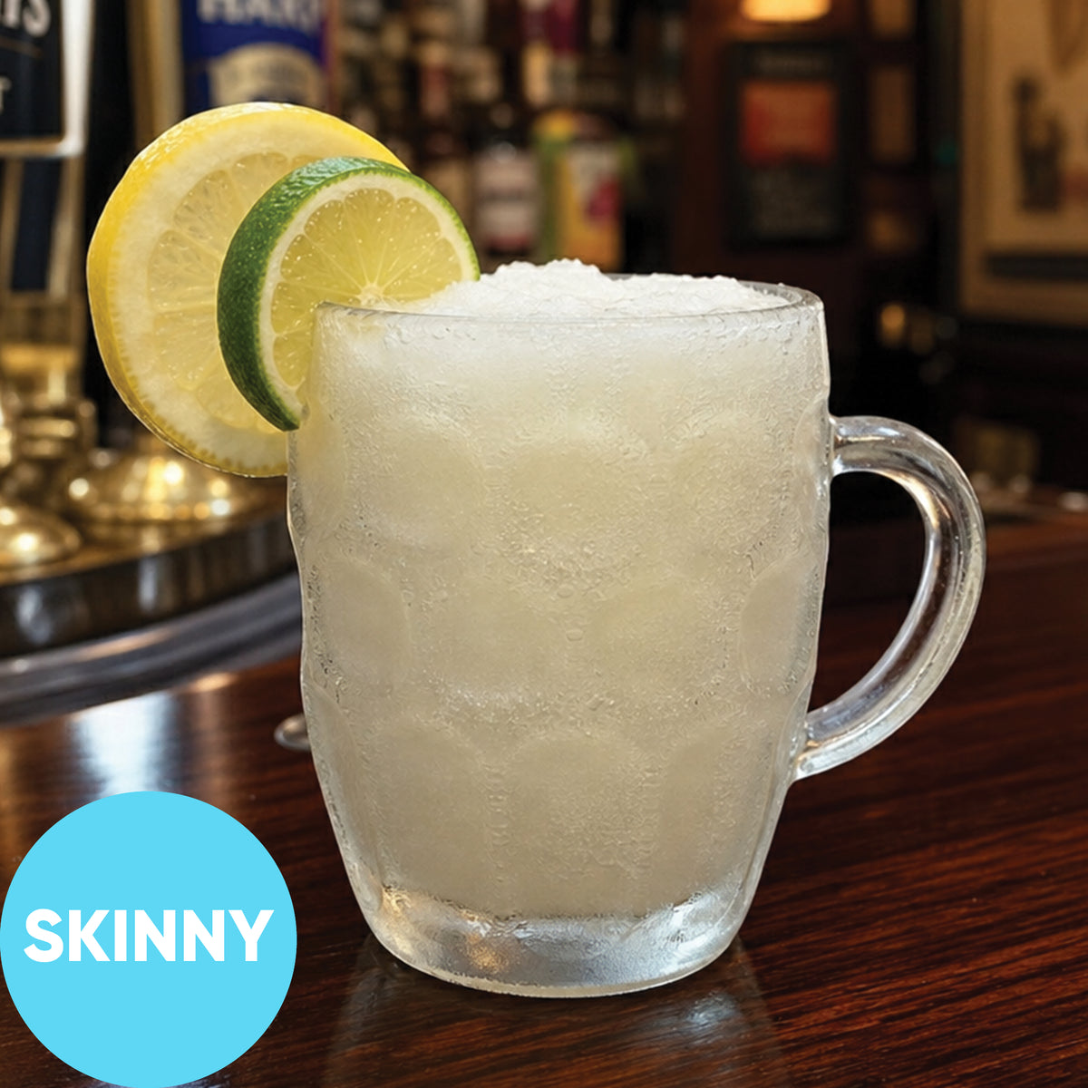 A frosted clear glass mug filled with Frozen Skinny Hard Lemon-Lime Pub Squash VINKA Slushy Kit (2L), garnished with two lime slices on the rim, set on a wooden bar counter with a blurred bar background. A blue circular badge in the lower left reads 'SKINNY'.