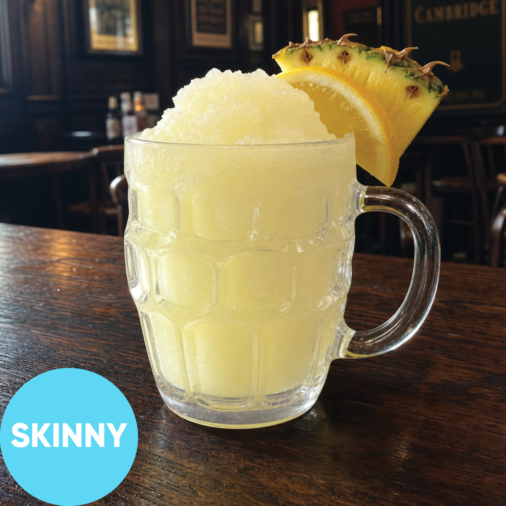 A clear textured glass mug filled with a Frozen Skinny Hard Lemon Pineapple Pub Squash VINKA Slushy Kit (2L), garnished with a pineapple slice, sitting on a dark wooden table. A blue circular badge in the lower left reads 'SKINNY'.
