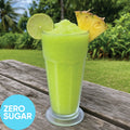A tall clear glass filled with Frozen Zero Sugar Hard Pine Lime VINKA Slushy Kit (2L), garnished with a lime wheel and pineapple slice, sitting on a weathered wooden table with a tropical garden background. A blue circle in the lower left reads 'ZERO SUGAR'.