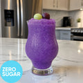 Frozen Zero Sugar Hard Purple Grape VINKA Slushy Kit (2L) in a clear stemmed glass on a white marble kitchen counter, with two grapes on top and a blue circle in the lower left that reads 'ZERO SUGAR'. A blurred kitchen and refrigerator are in the background.
