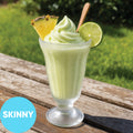 A Frozen Skinny Hard Creamy Pine Lime VINKA Slushy Kit (2L) in a clear glass, topped with whipped green swirl, a pineapple wedge, and a lime slice, sitting on a wooden table outdoors with a grassy background. A blue circle in the lower left says 'SKINNY'.