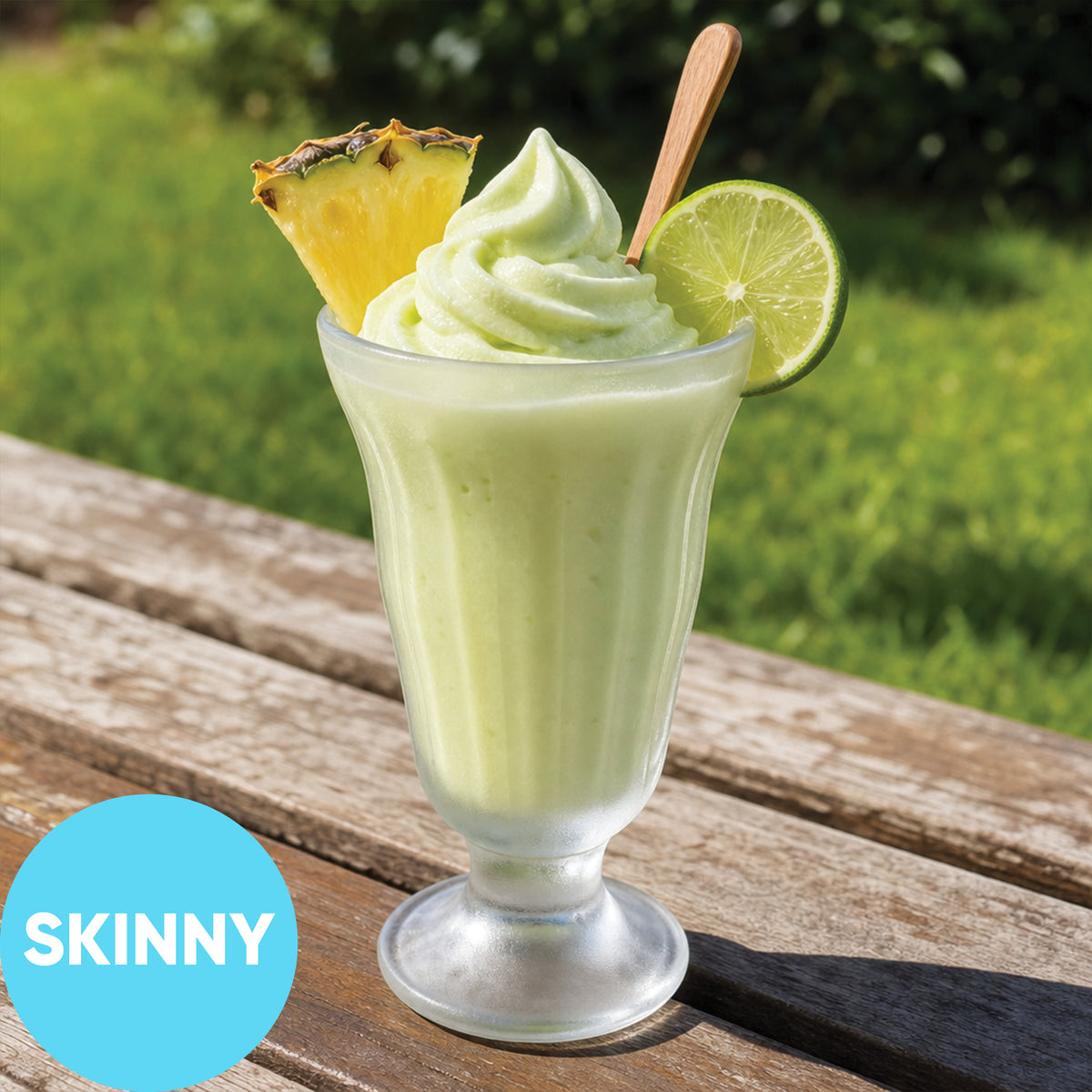 A Frozen Skinny Hard Creamy Pine Lime VINKA Slushy Kit (2L) in a clear glass, topped with whipped green swirl, a pineapple wedge, and a lime slice, sitting on a wooden table outdoors with a grassy background. A blue circle in the lower left says 'SKINNY'.