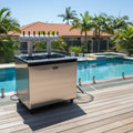 A stainless steel outdoor beverage dispenser cart with multiple beer taps, set on a wooden deck next to a swimming pool with palm trees and a house in the background.