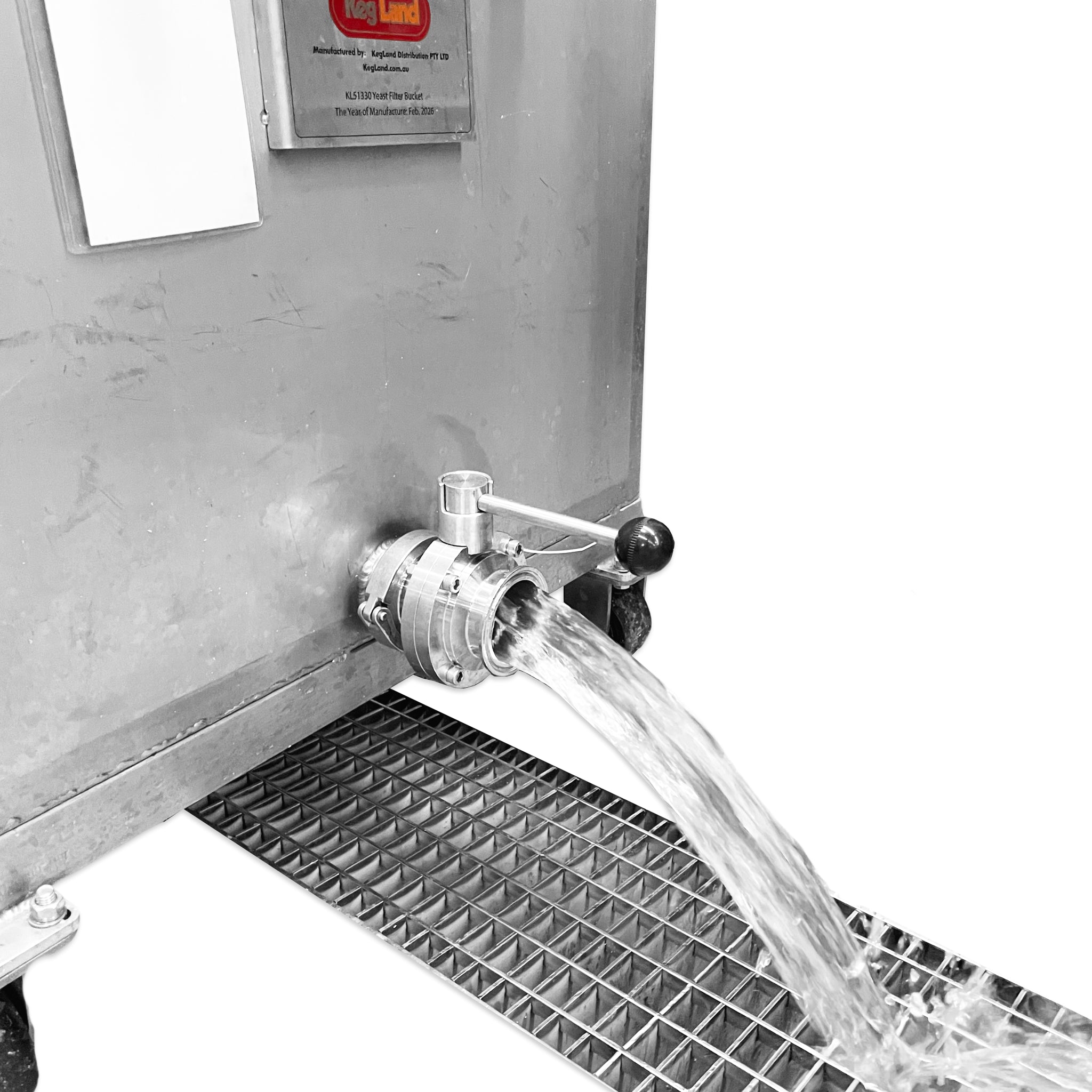 A metal outdoor water spigot with a manual lever valve, actively pouring water over a black metal drainage grate. A silver metal brand plate is visible on the wall above.